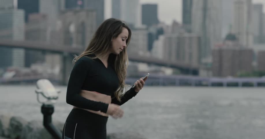 A woman holds her phone and stretches in front of the water and then runs off