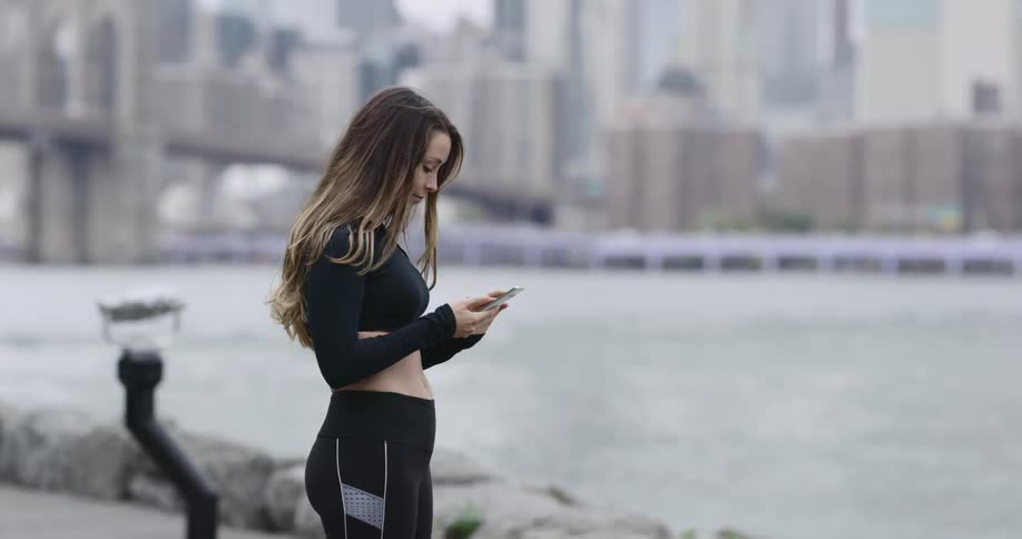 Woman in exercise clothing looks at her cellphone as she stands next to the river