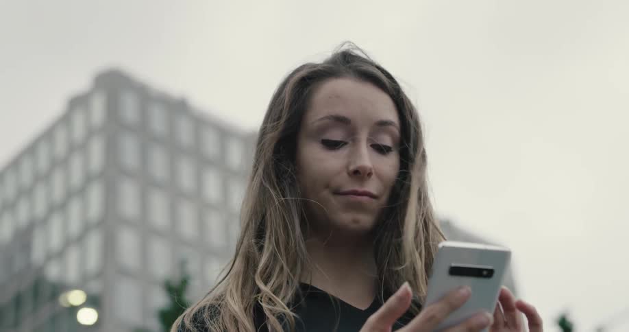 Woman wearing bluetooth earphones uses smartphone and looks around on a cloudy day in the city