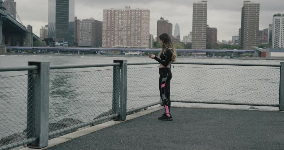 A woman stands on a pier beside the river looking at her cell phone then starts to run away