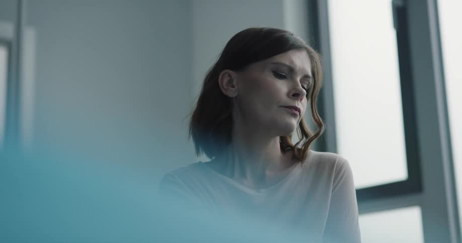 A woman sits in a clinic room and stares out the window with a contemplative look on her face