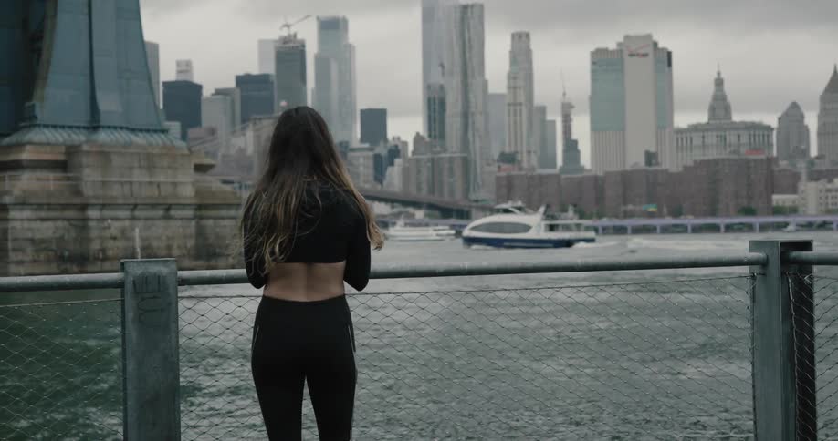 Woman wearing tights walks on the riverside and stops in front of the wire fence to check the smartphone in new york city on a cloudy