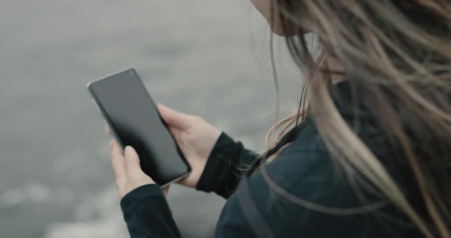 A woman puts in headphones and holds a smartphone while standing on a rocky beach
