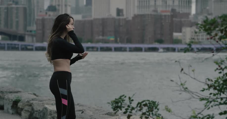 A woman runs up to a river bank and stretches her legs while holding her cell phone