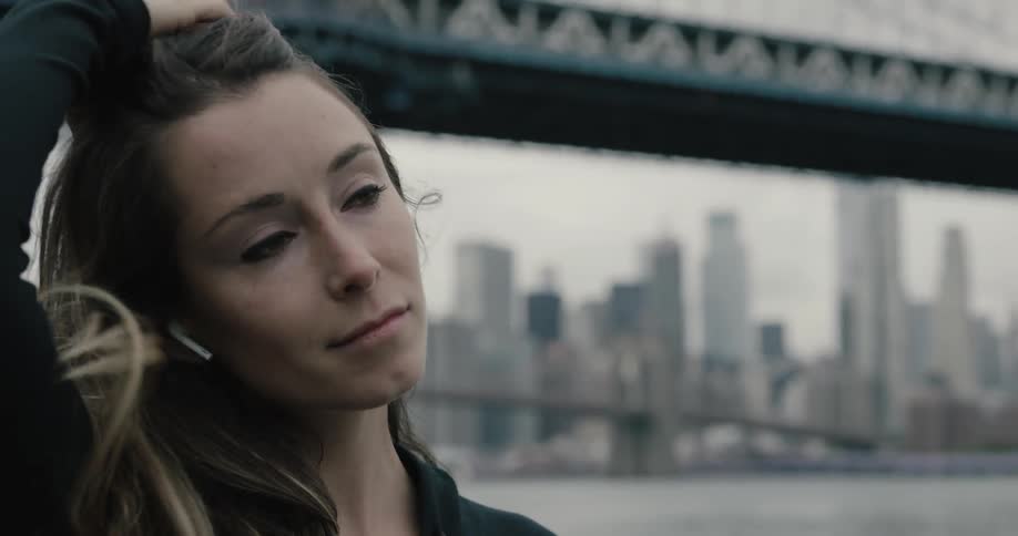 A woman stands beside a river fixes her earphones and tosses her hair