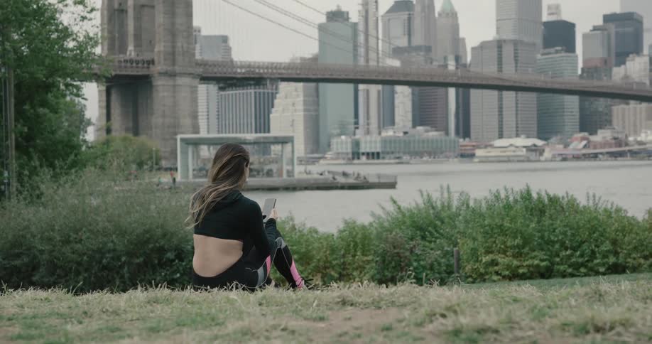Woman sitting on the grass using smartphone next to the river across the new york city and brooklyn bridge on a cloudy day