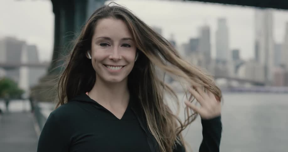 A woman smiles as she runs her hands through her hair as she stands by the edge of a river running through a metropolitan area