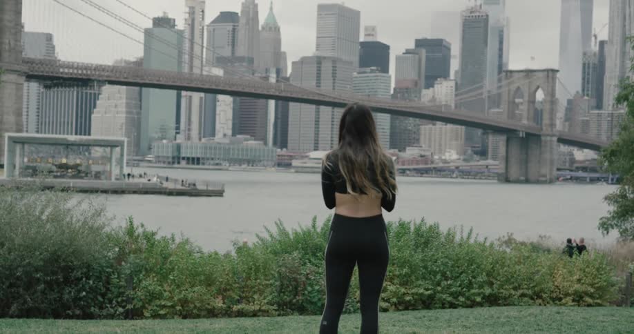 A woman wearing running clothes walks onto the grass and looks out at the city skyline harbor and bridge