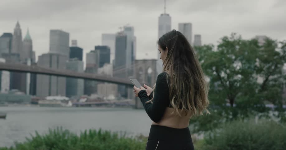 A woman in a crop top uses a smartphone in a park overlooking a city bridge and skyscrapers