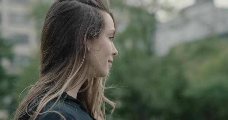 Woman wearing bluetooth earphones smiles and looks ahead on a cloudy day in the city