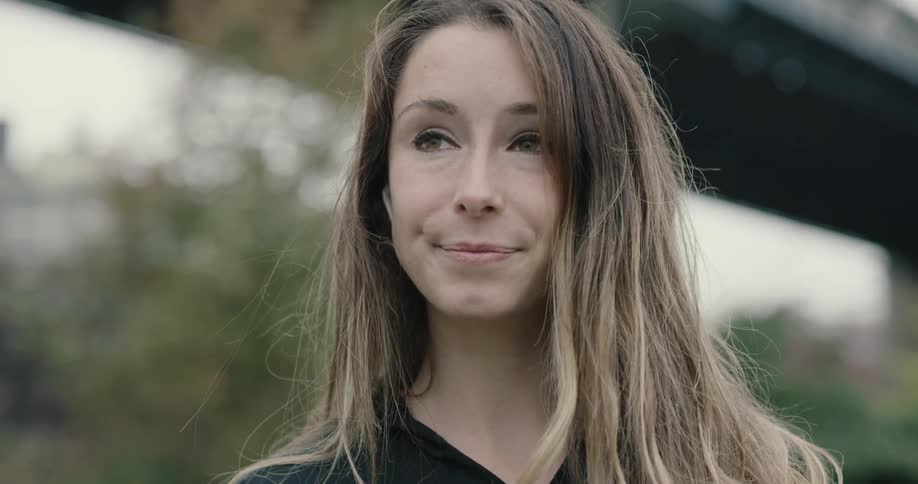 Woman wearing bluetooth earphones looks at the camera and smiles in the city on a cloudy day