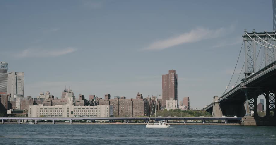 A boat drives under a bridge on a river surrounded by city buildings and roadways