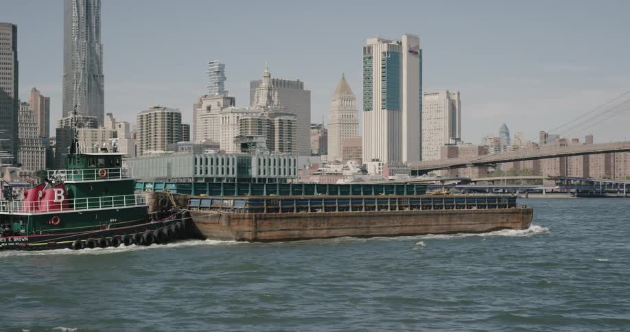A tugboat pushes a barge over the water as it passes by a major metropolitan city and heads towards a bridge that spans the body of water