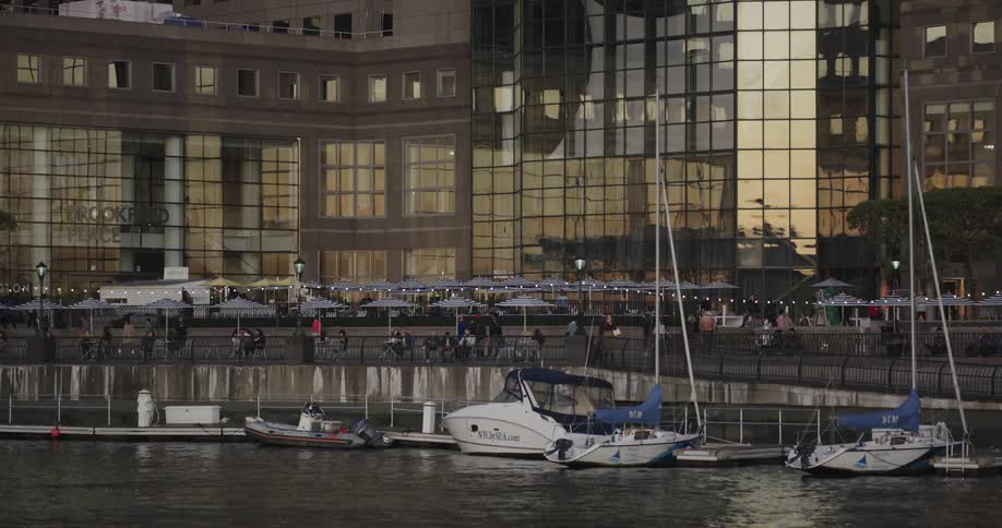Several small boats are docked along side a dock next to a marina full of people sitting at tables and walking