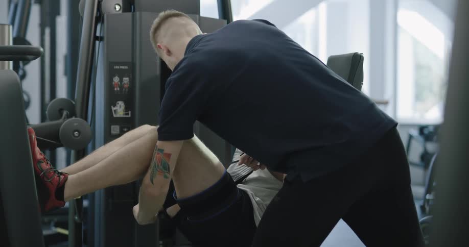 A young man sits on a leg press machine while another man puts a wrap on his leg