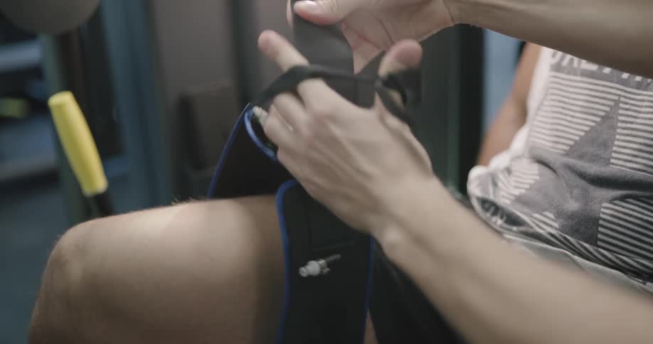 A man places a strap around the thigh of another man who is seated in an apparatus within a physical therapy gym
