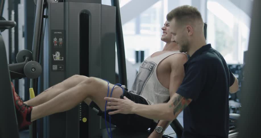 One man working out on a leg machine while the other man trains him