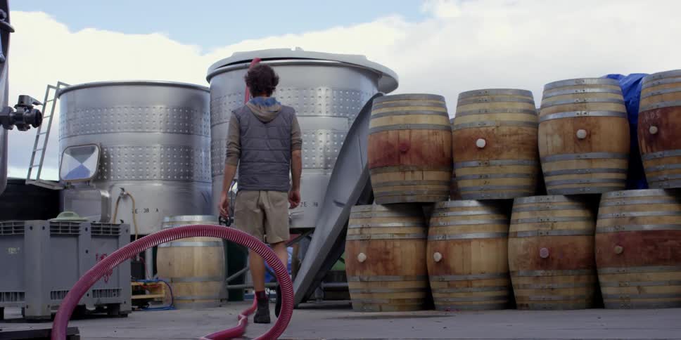 A man walks past a stack of barrels toward large metal containers