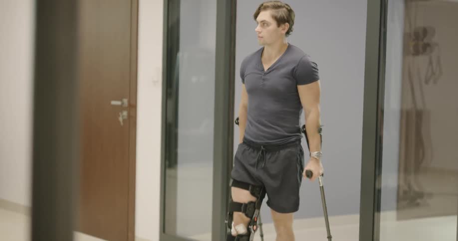 A man in a leg brace and using crutches leaving a room and being shown down the hall by another man