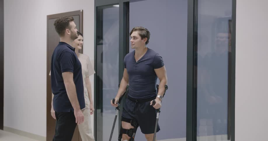 A man walks to a door where a woman and a man with a leg brace and crutches come out