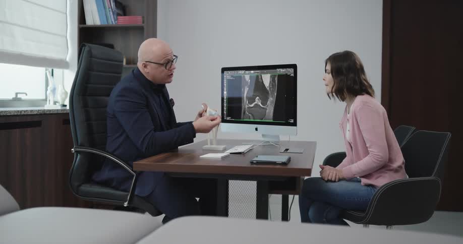 A male medical professional holds a model of a knee joint on his desk as he shares information with a female patient