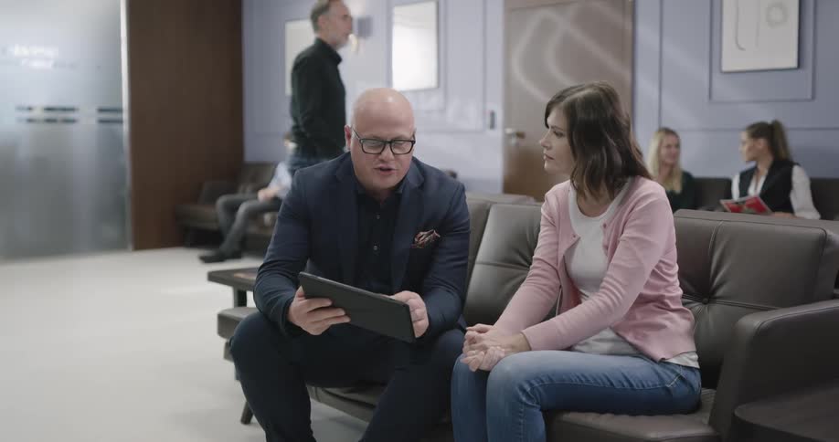 A bald man in a suit coat sits with a female patient in the waiting area of a clinic and discusses information that he is showing her on a handheld electric tablet
