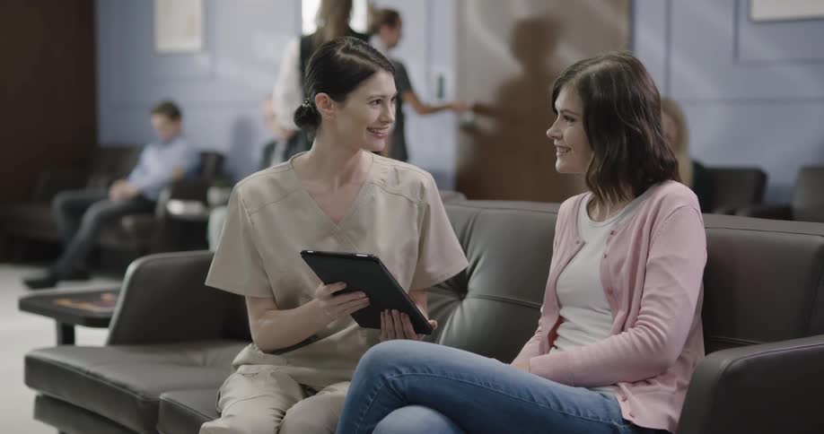 A medical professional speaks with a smiling woman while holding a tablet in a waiting room
