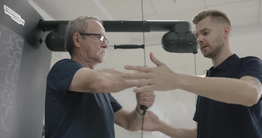 A younger man helps an older man who is performing an exercise in which he extends his arms while holding a pulley that is fixed to a piece of exercise equipment