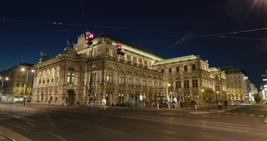 A building with elaborate architecture stands at the corner of a very busy intersection at night as people cars and trolleys pass by at night