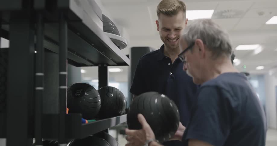 A young man picks up a medicine ball and hands it to an elderly man