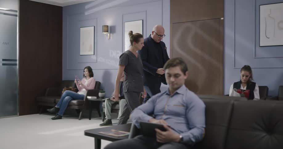 A man walks through a waiting room with a nurse in scrubs and talks as he shows her information on a tablet