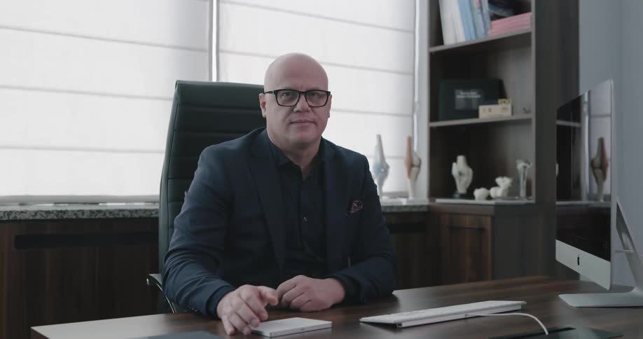 A bald man sitting at a desk using an apple computer before turning to look at the camera