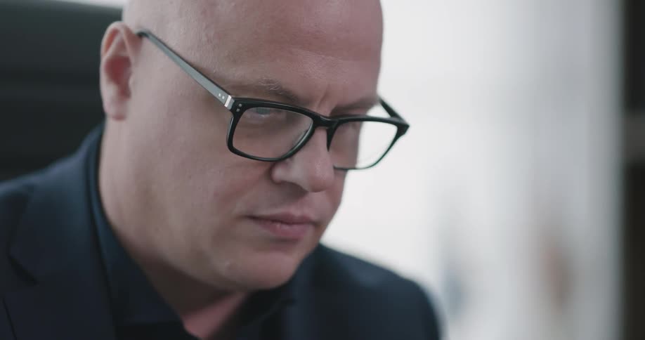 A bald white man with glasses sits in a sunlit room