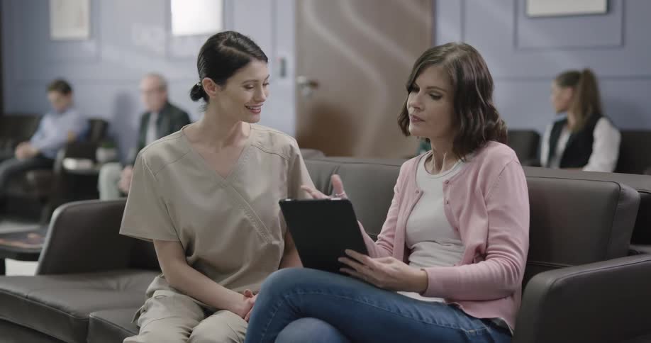 A nurse sitting with and talking to a woman on a sofa in a lobby while looking at a tablet