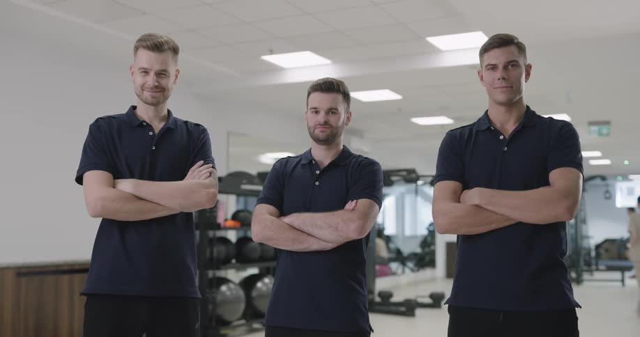 Three men stand side by side with their arms folded while smiling confidently as they stand in front of a fitness room filled with equipment
