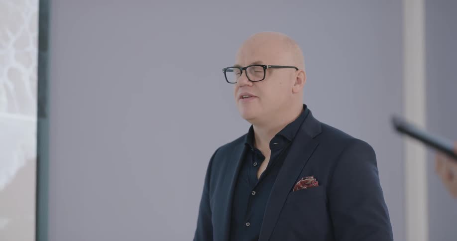 A bald man is talking in front of a screen with a presentation on it