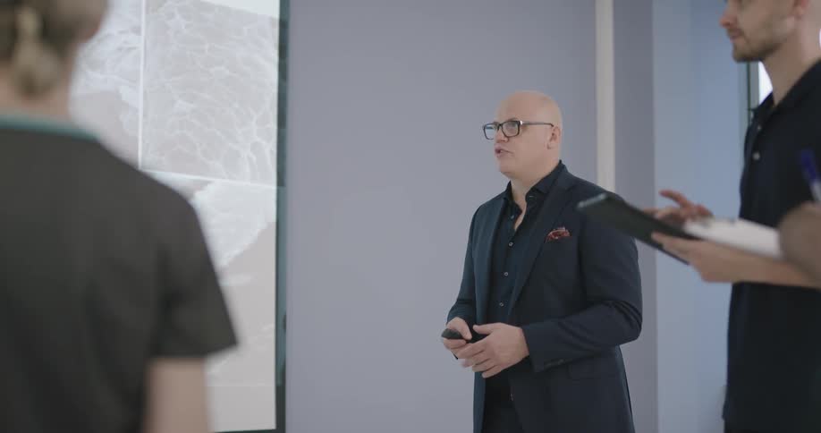A group of people holding tablets stand and listen to another man give a presentation