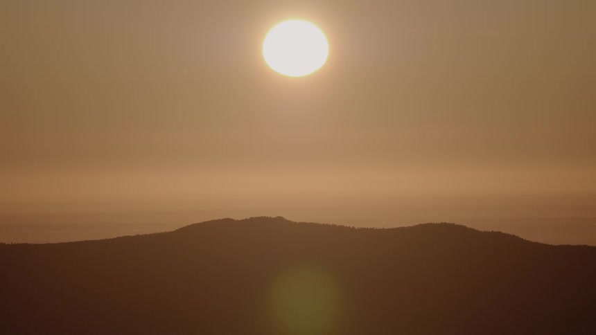 The sun hangs low in the sky above a forested mountain range on a hazy day leading to an orange tinted sky