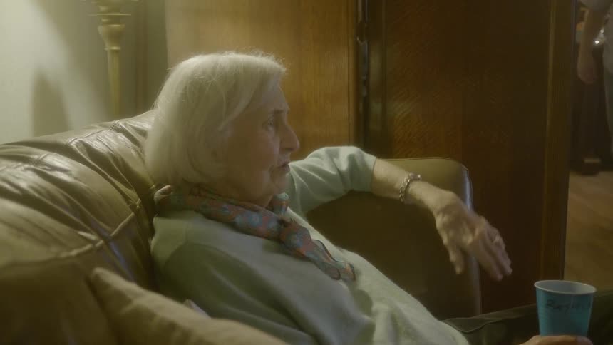 An elderly woman sits on a leather sofa while holding a plastic cup