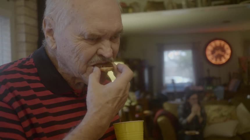Old man eats cookie and finds it tasty in the living room with people sitting on the couch on the background during daylight