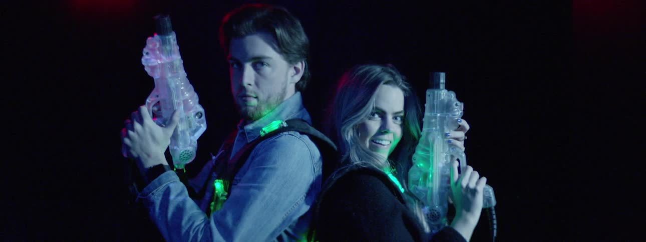 A man and woman playfully stand back to back while wearing vests with flashing lights and holding toy blasters