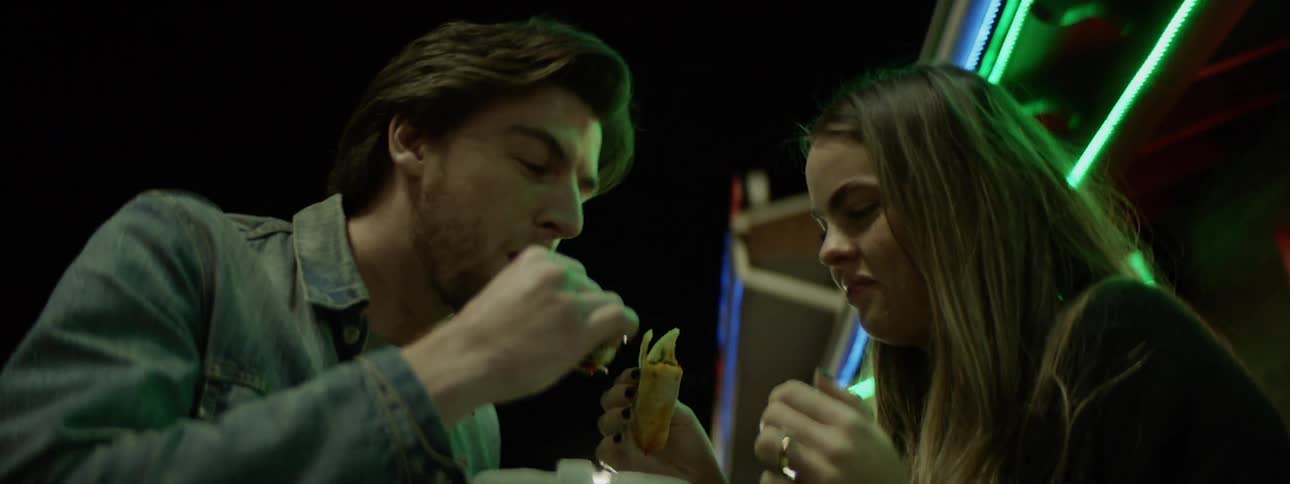 A man and woman eating food from a takeout container under neon awning lights