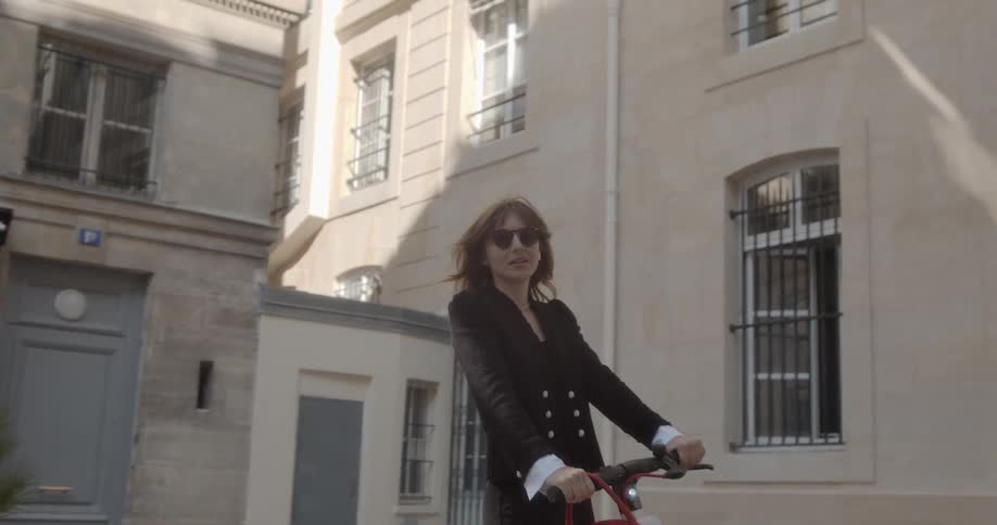 A woman rides a scooter next to some tan buildings