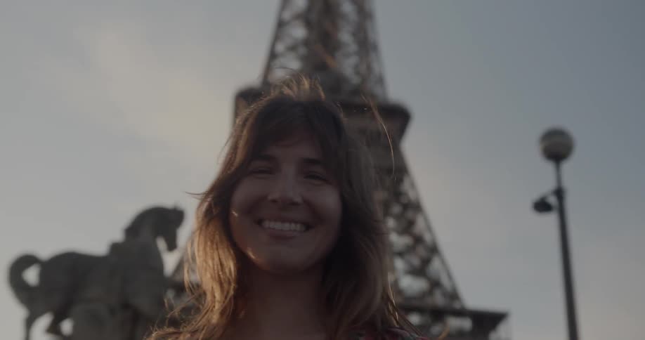A woman with brown hair in a white shirt with red flowers stands beside the eiffel tower in champ de mars park under a sunset