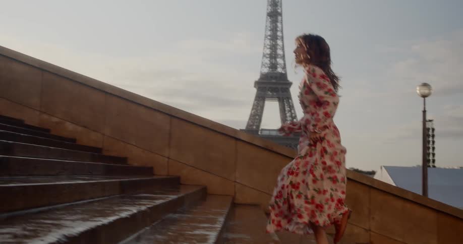 A brunette woman in a long dress runs up a set of stairs with the eiffel tower visible in the background