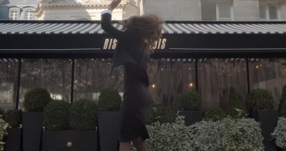 A woman wearing sunglasses and a dress with a suit coat dances and spins around in front of a bistro