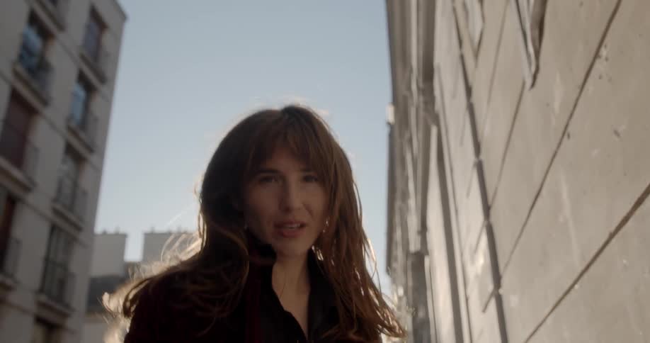A woman walks backwards and stares into the camera in an urban setting