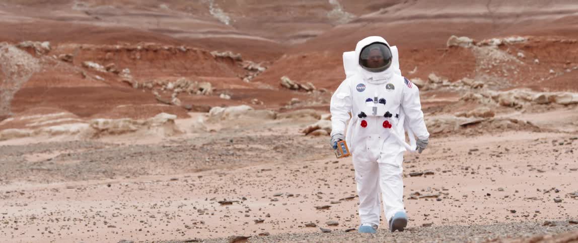 A person in a space suit walks across rocky soil