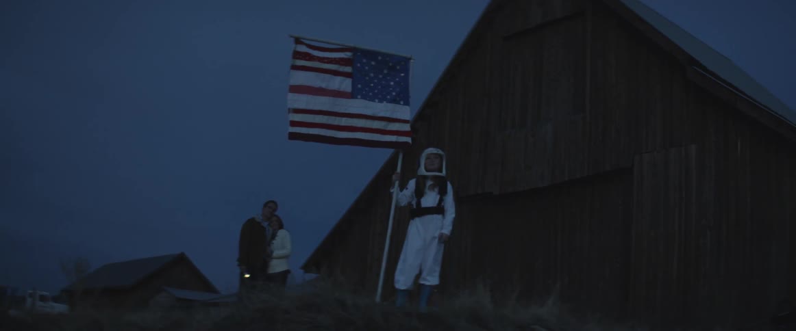 A person in an astronaut costume planting a fake american flag into the ground outside of a wooden barn