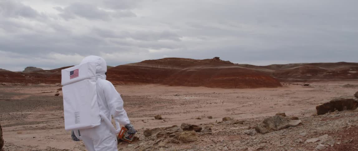 A person wearing an astronaut costume walks along a barren landscape that resembles the desolate surface of an uninhabited planet